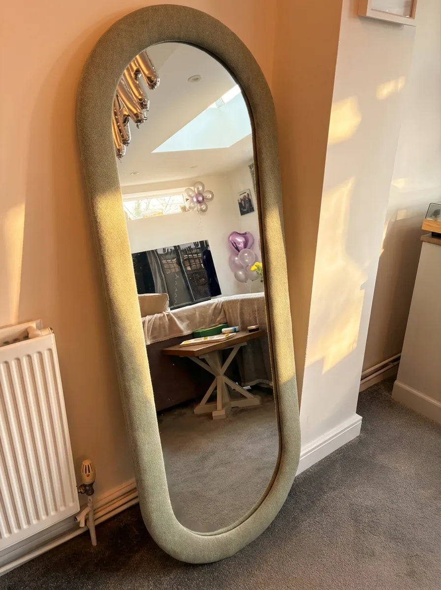 Oval mirror with decorative fabric frame leaning against a wall in a room.