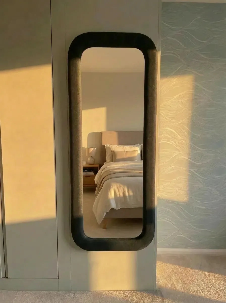Upholstered Fabric Rectangular mirror on a wall reflecting a bedroom with a bed and pillows.